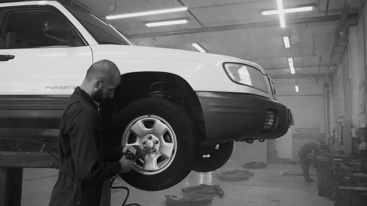 A mechanic crouched at the front wheel of a car in a workshop bay.